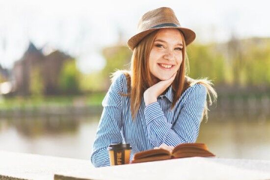 Woman Smiling Reading the Bible A girl wearing a hat and striped shirt sits at a bench near a lake with her Bible. One way to spend Sabbath is out in nature and in God's Word.
