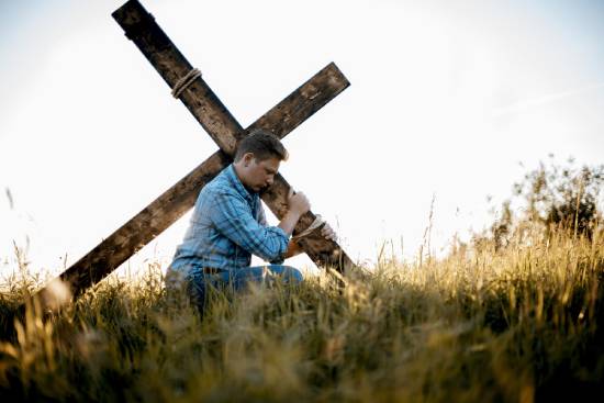 A man carries a cross over his shoulder as he walks through a field, illustrating Jesus' teaching about taking up our crosses (Mark 8:34).