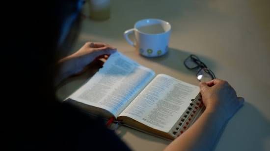 Woman reading her Bible at a table with a cup of tea A woman reads her Bible with a mug and glasses nearby. This illustrates studying God's Word and taking it to heart.