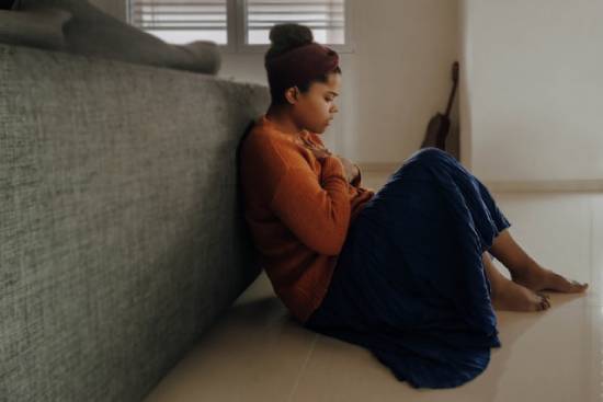 A woman sitting on the couch a young woman of color who looks distraught sits on the floor leaning against the back of a couch with a blanket over her legs