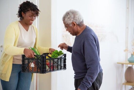 A young woman happily offers a basket of produce to an elderly man.