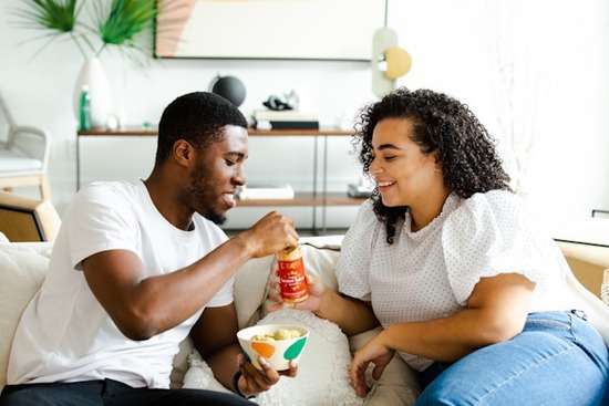 Man and Woman Living Together Before Marriage A cohabiting couple trade snacks as they sit together on their living room couch.