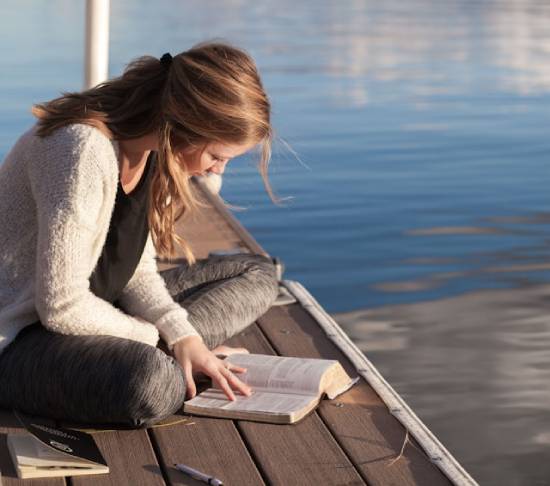 A woman sitting on a dock reading her Bible demonstrates how the best way to avoid legalism is the spend personal time with God, and get to know our Savior through the Bible.