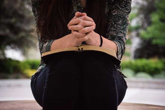 Woman praying with her Bible open on her lap Woman sitting while praying with folded hands on an open Bible as we learn about various physical postures during prayer