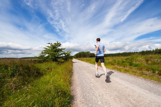 A man in activewear runs on a dirt road by a grassy field.