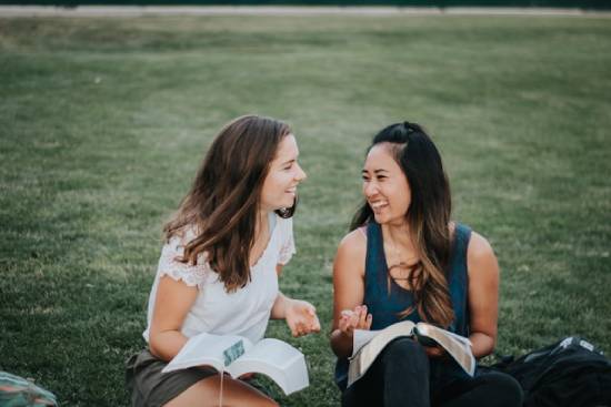 Two young women joyfully study the open Bibles on their laps as they sit on a lawn.
