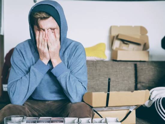 A young man in a hoody sits on a couch with his head in his hands, distraught about something.