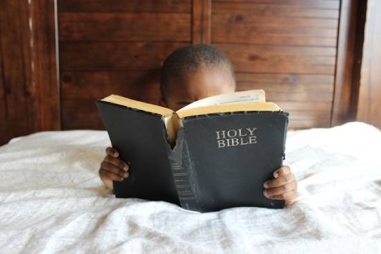Young boy reading a large Bible while lying on a bed
